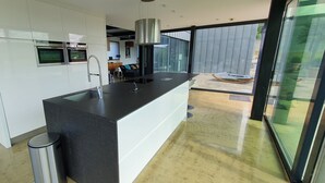A modern kitchen with built-in ovens, a large island with a sink, and a view of the outdoor patio through large windows. - VILLA COMBINES MODERNITY AND STONES. IDEAL FOR FAMILY OR WORK (LIFFRE)
