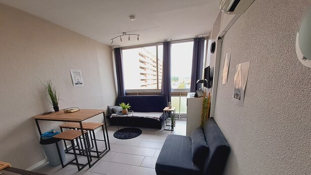 A compact living space with a kitchenette, a dining area, a sofa, and a view of a building through large windows. - Lovely studio overlooking the Alberts at the port, with the sea in front (Saint-Cyprien)
