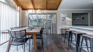 Un comedor moderno con una mesa de madera y sillas negras, una zona de bar con taburetes, y vistas a través de un vidrio.