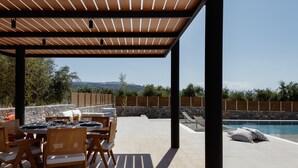 En udendørs patio med et pergolaoverdækket område, spisebord, stole og en swimmingpool.