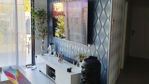 A modern living room with a large TV mounted on a patterned wall, a white entertainment unit, a colorful rug, and a black speaker. - VILLA WITH JACUZZI (cers)