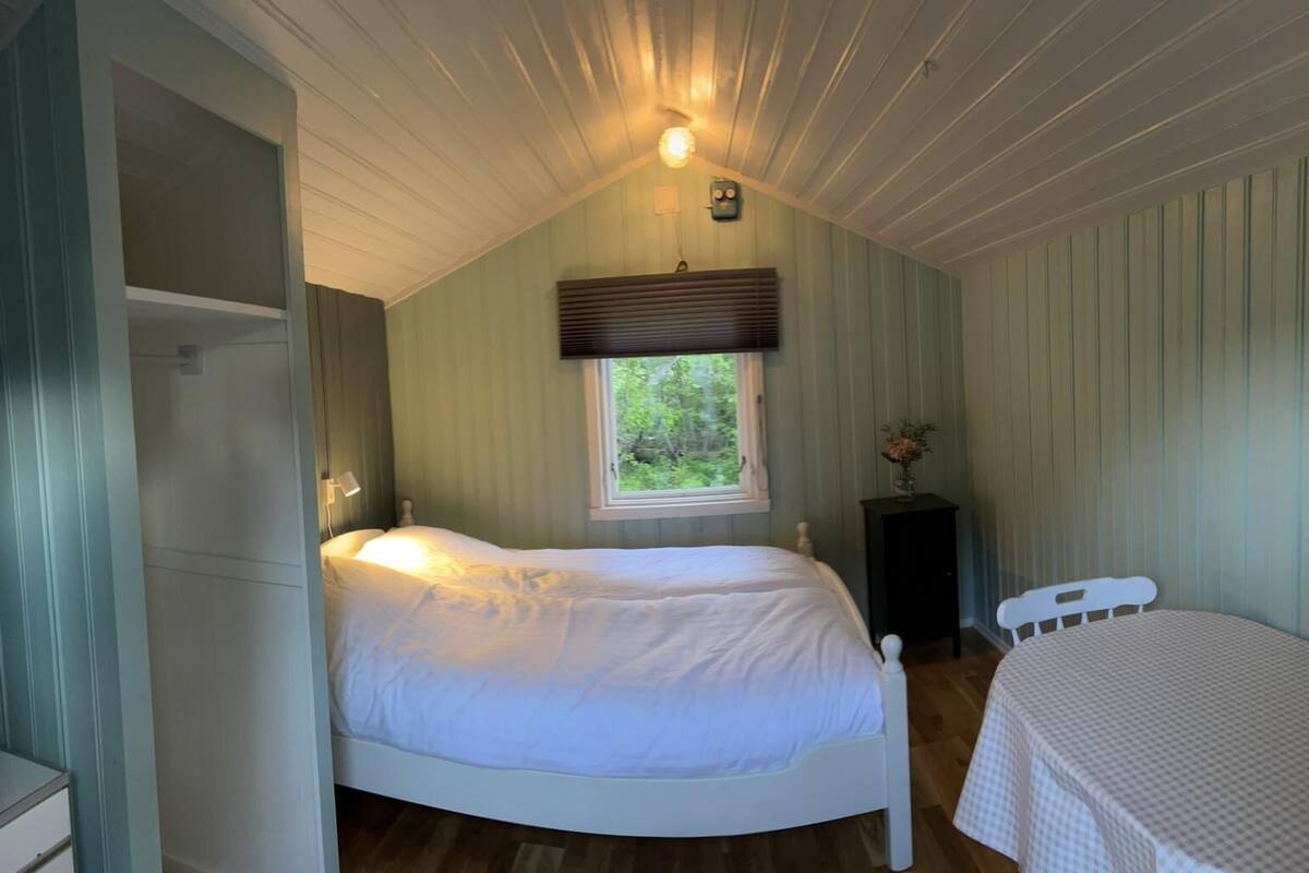 A bedroom with a single bed, a small bedside table with a vase, a window with blinds, and a door leading to another room.