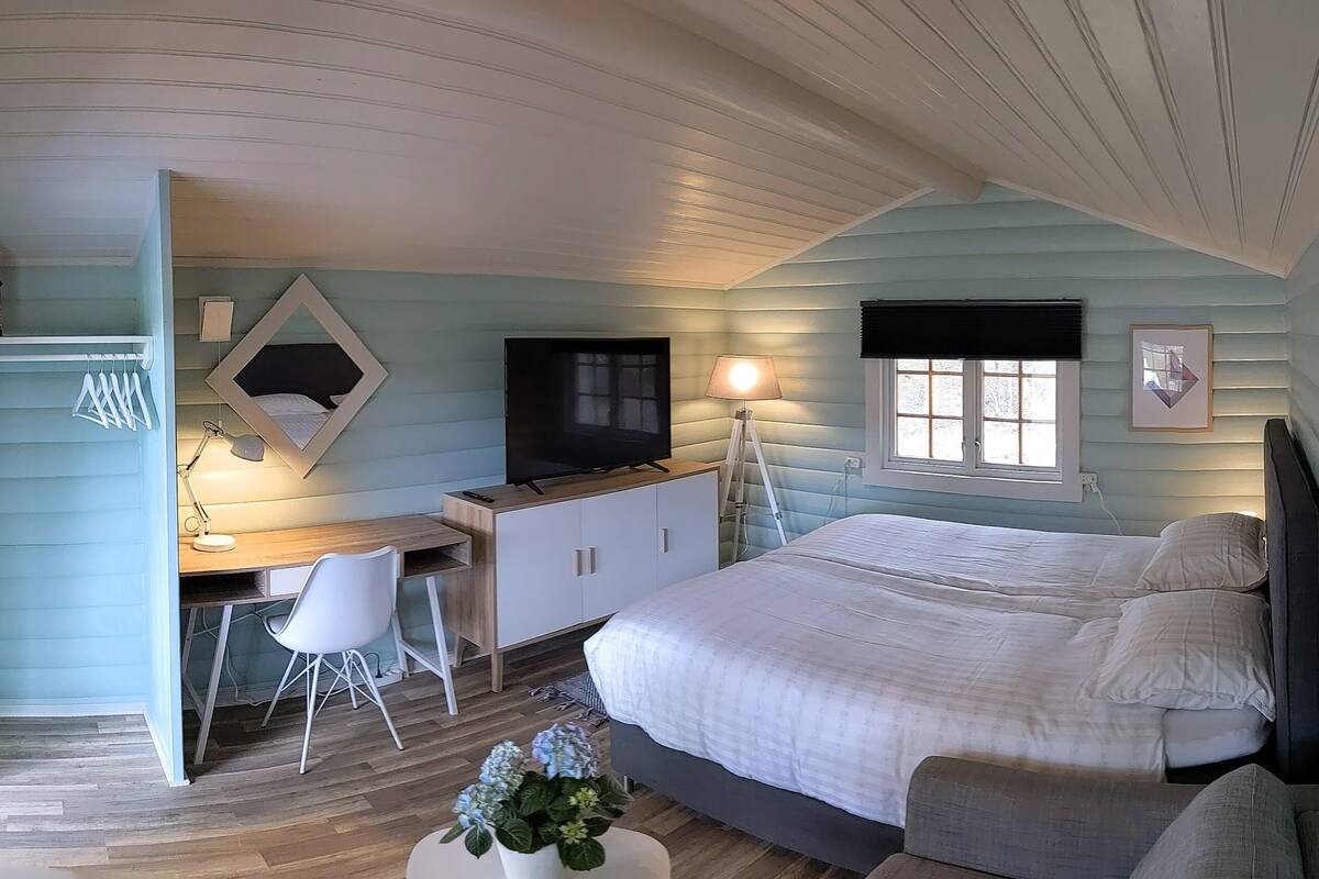 A bedroom with a bed, desk, chair, TV, lamp, window, and artwork.