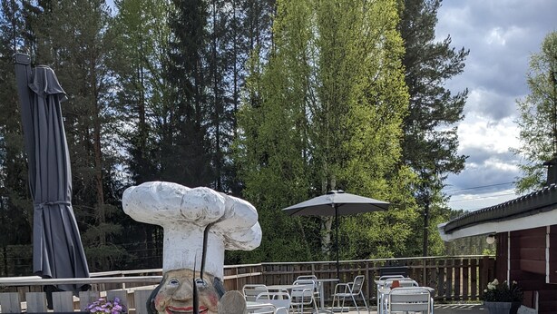 A wooden deck with a statue of a chef, outdoor tables, chairs, and a flag. - Neset Skydsstasjon (Flesberg)
