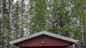 A small, red cabin with a porch and a single window, surrounded by a forest. - Neset Skydsstasjon (Flesberg)