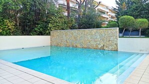 Una piscina con muretto in pietra e pavimento piastrellato, circondata dal verde e da un edificio sullo sfondo.