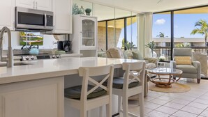 Cozinha moderna com armários brancos, micro-ondas embutido e fogão de aço inoxidável. Possui área de jantar com cadeiras brancas e mesa de vidro. O ambiente tem grandes janelas com vista para palmeiras e um edifício.