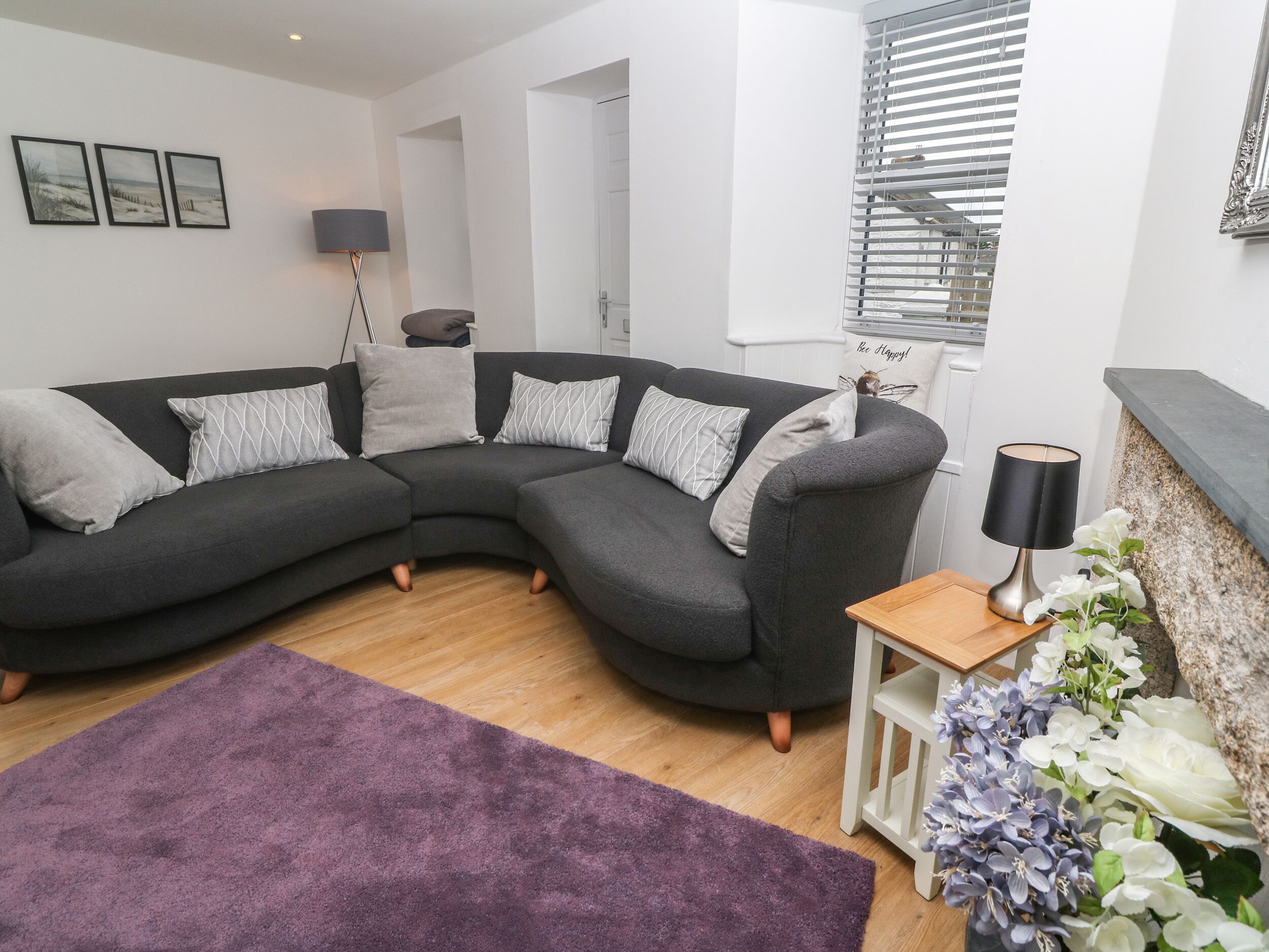 Ein modernes Wohnzimmer mit einem dunkelgrauen Ecksofa, einem violetten Teppich, einem kleinen Holznachttisch mit Lampe und einer Zimmerpflanze.