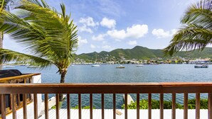 Una terraza de madera con vistas a un cuerpo de agua tranquilo, con barcos, un pueblo lejano y montañas.