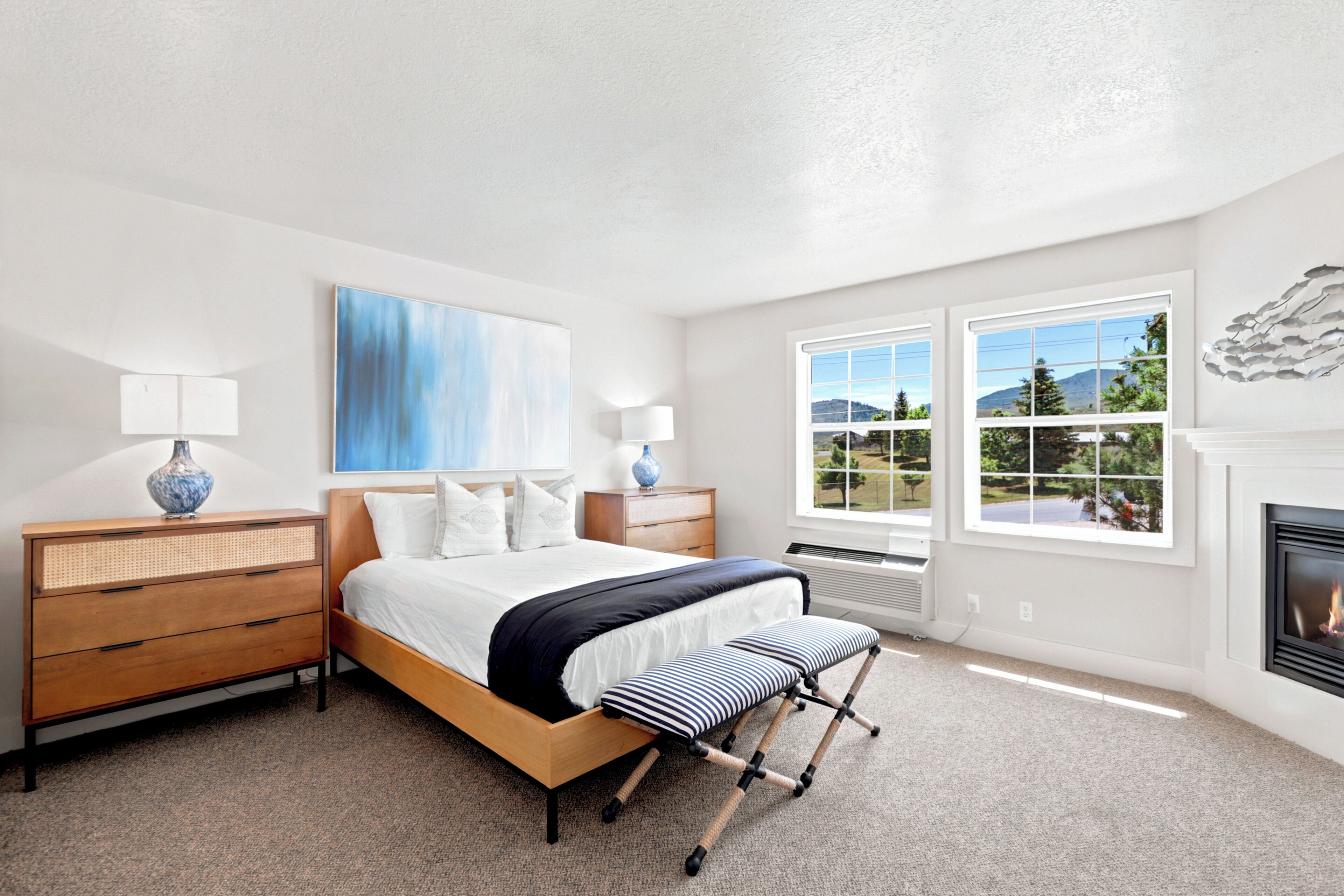 A bedroom with a bed, bedside tables, a lamp, a painting, a fireplace, and a view of the outdoors.