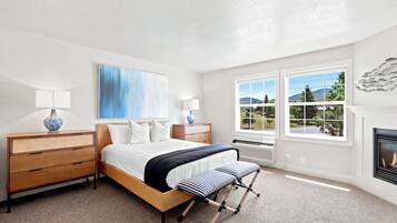 A bedroom with a bed, bedside tables, a lamp, a painting, a fireplace, and a view of the outdoors.