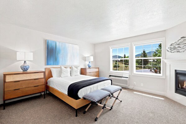 A bedroom with a bed, bedside tables, a lamp, a painting, a fireplace, and a view of the outdoors.
