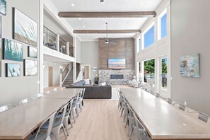 A spacious dining area with a long table and chairs, a kitchen with a stone backsplash, and a staircase leading to an upper level.