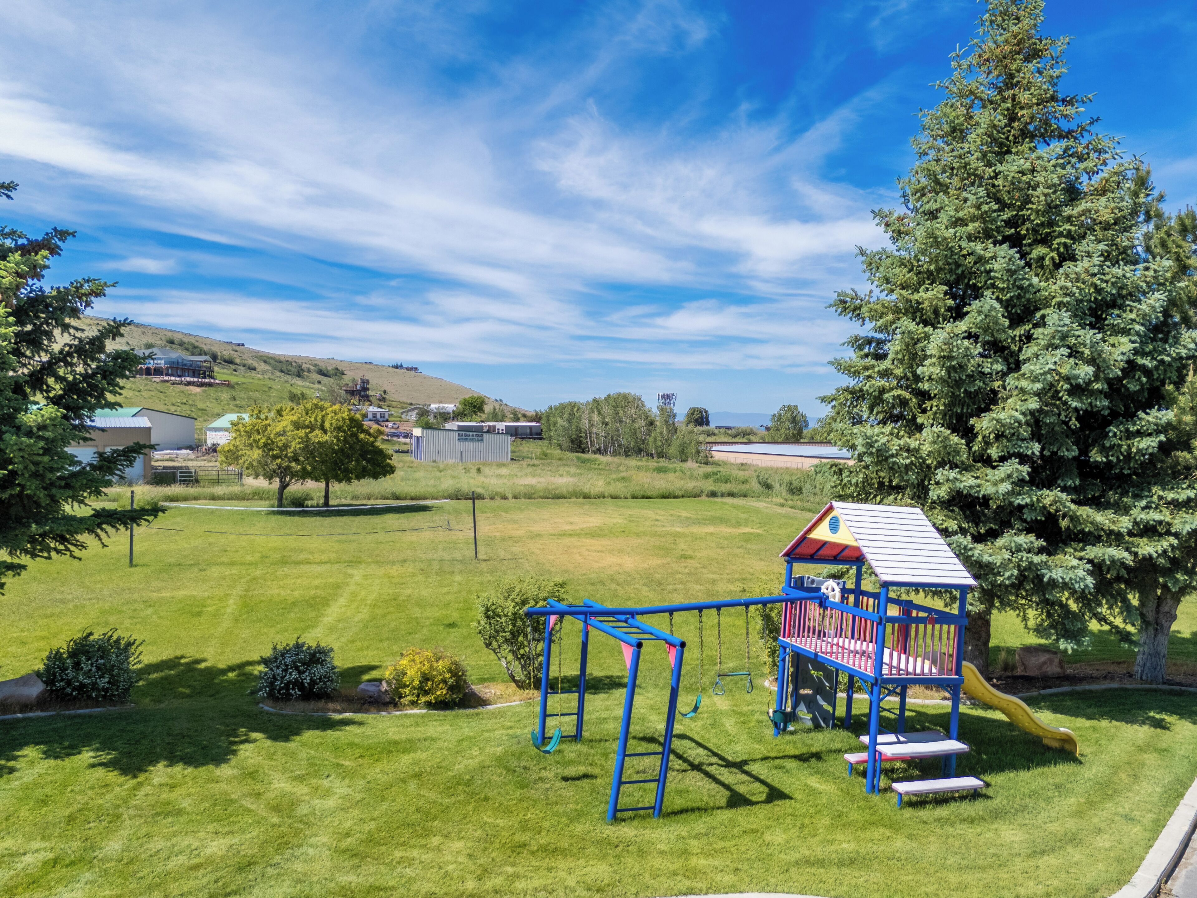 A playground with a slide and swings, surrounded by grass and trees, with buildings in the background.