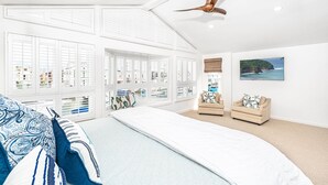 Un dormitorio con una cama grande, dos sillones, un ventilador de techo y vistas al mar a través de amplios ventanales.