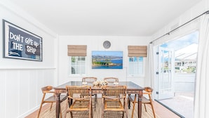 Un comedor con una mesa grande y seis sillas de mimbre, un reloj de pared y un cuadro enmarcado con un paisaje costero.