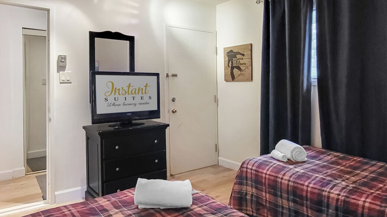 Une chambre d’hôtel avec deux lits, un téléviseur posé sur une commode, un miroir, une porte et une fenêtre avec des rideaux.