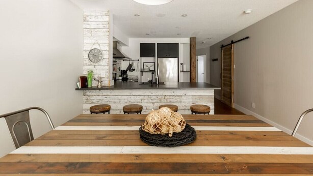 Une salle à manger moderne avec une table en bois et des tabourets de bar, une cuisine en arrière-plan et une horloge murale au style rustique.