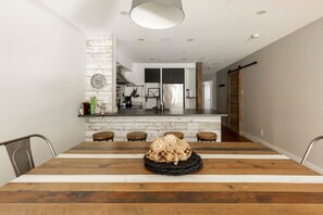 Ein moderner Essbereich mit Holztisch und Barhocker, im Hintergrund eine Küche und eine rustikale Wanduhr.