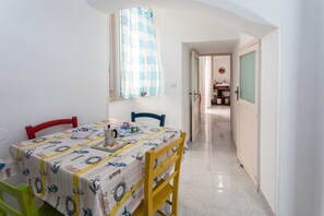 A dining area with a table set for two, a window with a checkered curtain, and a hallway leading to a kitchen area.