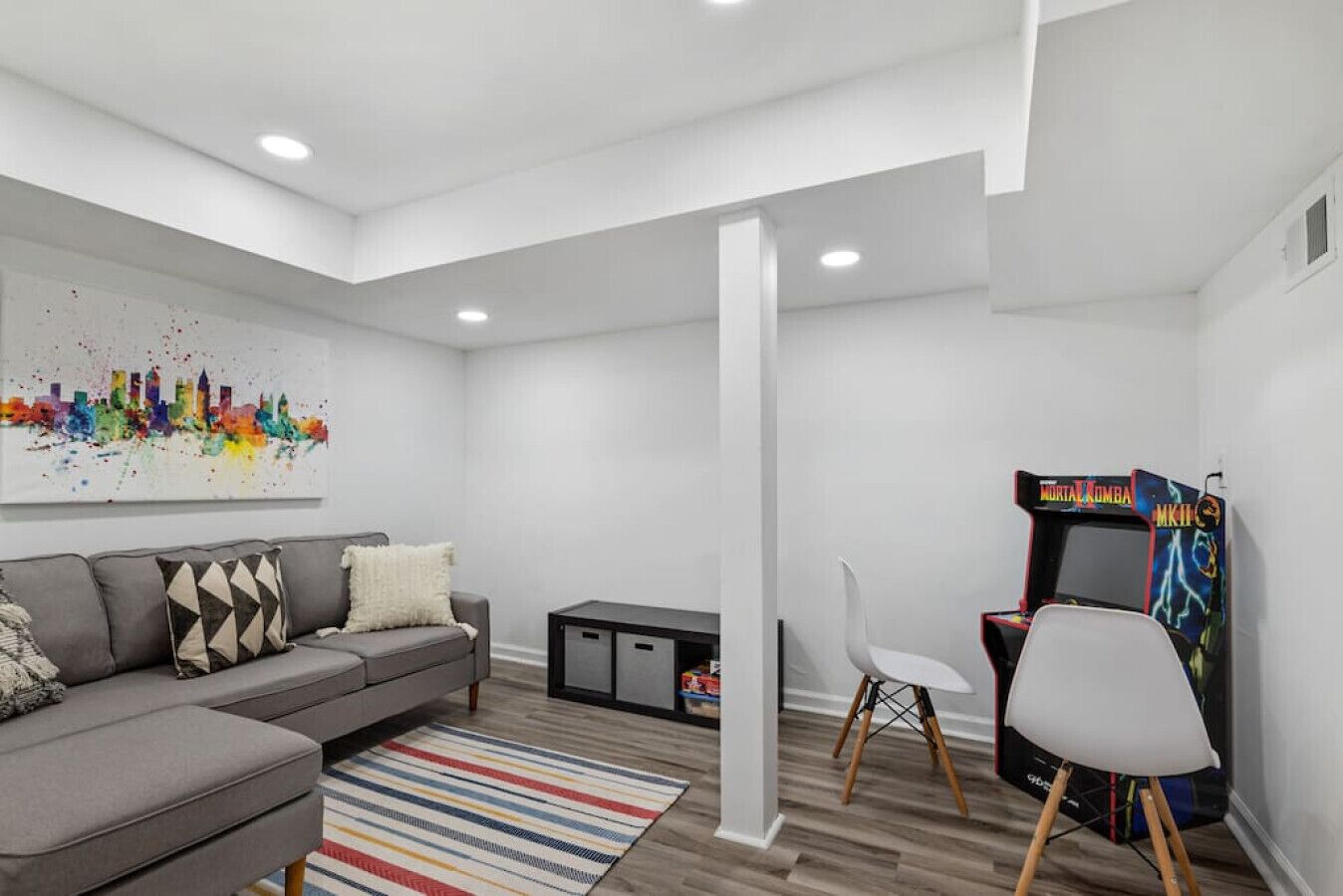 Un salon moderne avec un canapé gris, un tableau représentant un paysage urbain aux couleurs vives, un tapis à rayures et une borne d’arcade du jeu Mortal Kombat.