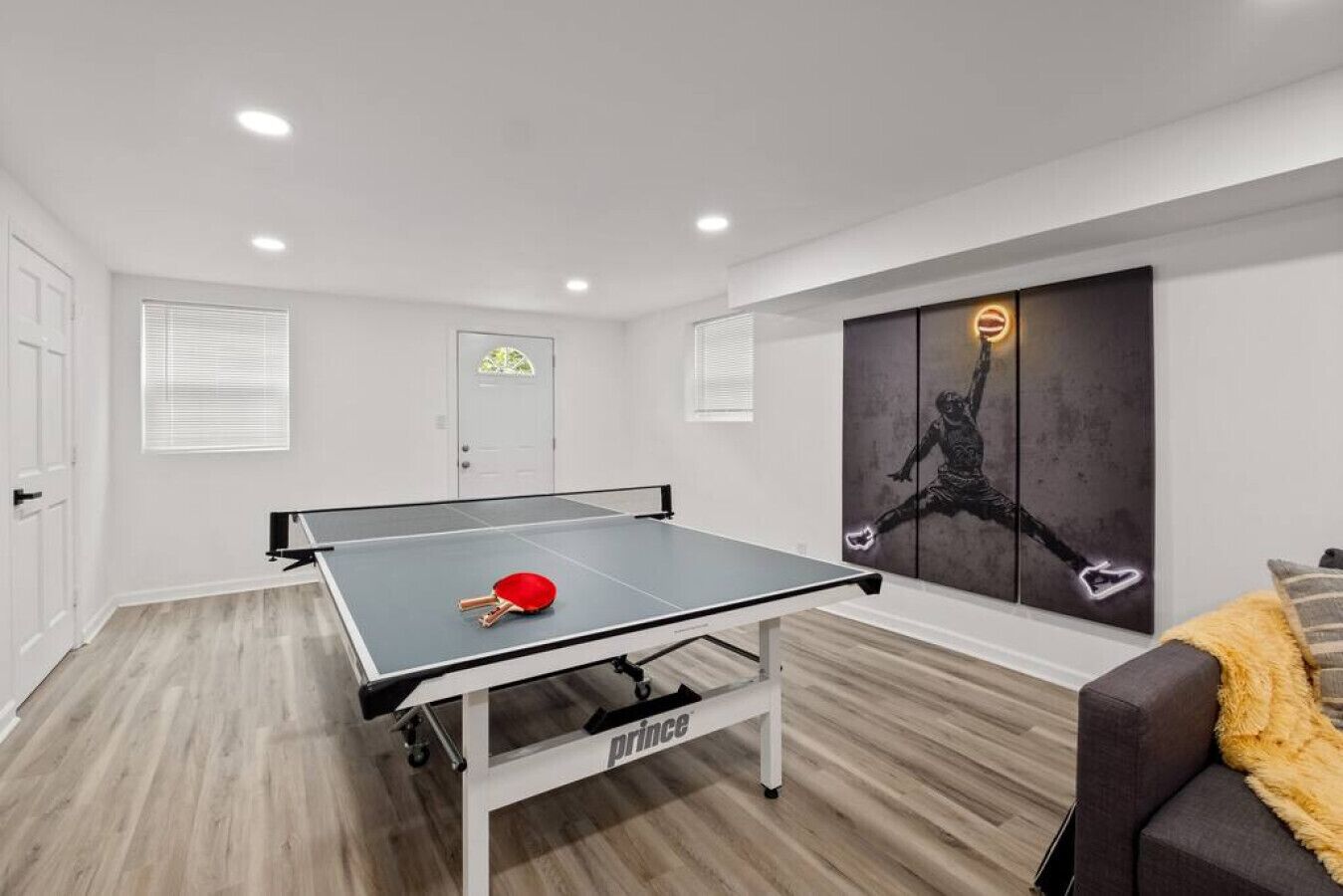 Taula de ping-pong amb una paleta vermella, un sofà gris i una obra d'art mural d'un jugador de bàsquet.