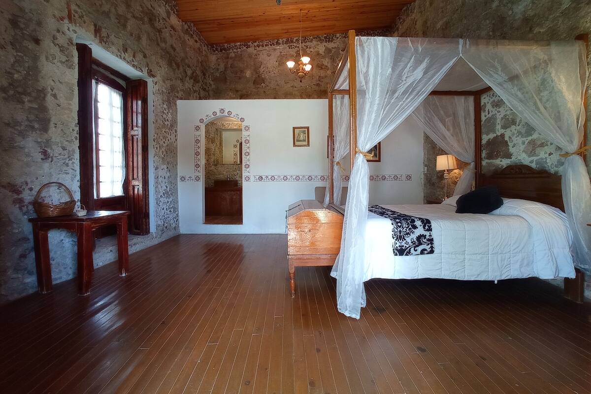 Une chambre avec un plancher en bois, un lit à baldaquin, un miroir et un banc en bois.