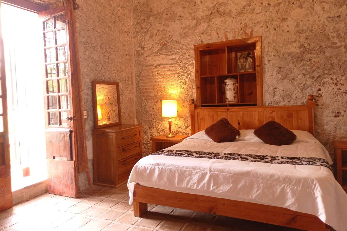 Une chambre à coucher rustique avec un lit en bois, une table de chevet avec une lampe, une commode et un mur en pierre.
