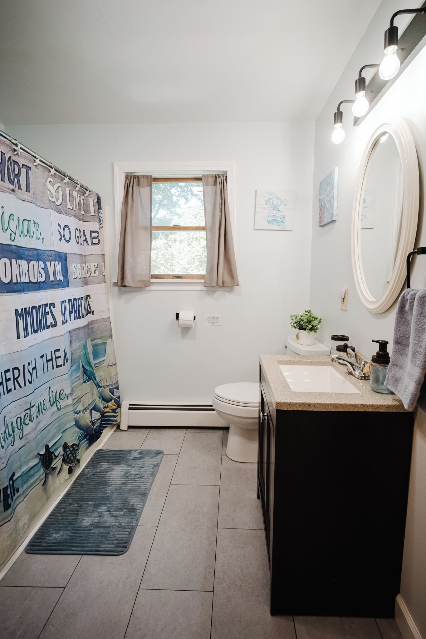 A bathroom with a shower curtain featuring motivational quotes, a window with curtains, a sink with a mirror, and a towel rack.