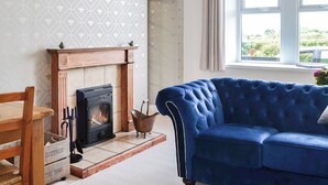 A cozy living room with a fireplace, a blue sofa, and a window with a view of greenery. - Bogrie Country Cottage (Annan, Dumfries)