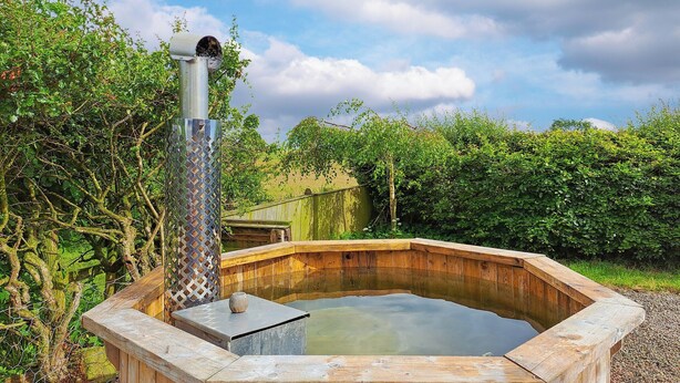 A wooden hot tub with a metal chimney, surrounded by greenery and a wooden fence. - Bogrie Country Cottage (Annan, Dumfries)