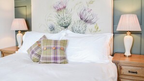 A neatly made bed with white linens, a plaid pillow, and a floral headboard. There are two bedside tables with lamps and a wooden nightstand with drawers. - Bogrie Country Cottage (Annan, Dumfries)