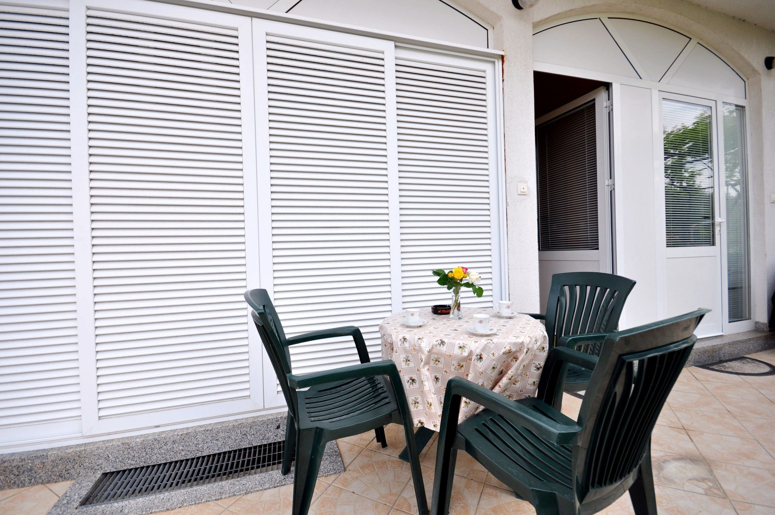 A patio with a table and chairs, a flower vase, and a closed white door with horizontal blinds.