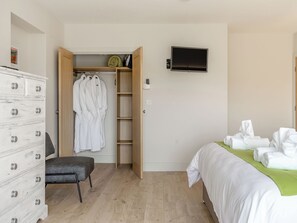 Ett sovrum med en byrå i trä, en stol, en säng med vita sängkläder, en grön pläd och en tv som är monterad på väggen.
