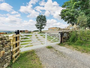 A rural property with a gravel path leading to a white gate, stone walls, and a large tree.