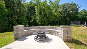 A circular outdoor seating area with a fire pit, surrounded by a brick wall, set in a grassy area with trees in the background. - Stunning 5 BD Home away from Home! 15 min to uptown (Charlotte)