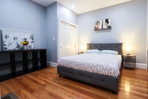 Ein Schlafzimmer mit einem grau gepolsterten Bett, zwei Nachttischlampen, einer weißen Tür und einer schwarzen Regaleinheit mit Blumen in einer Vase.