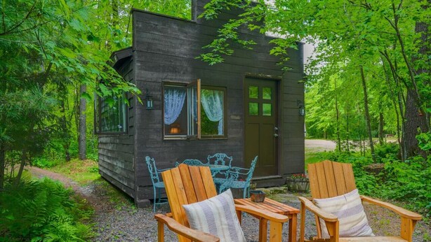 Une cabane avec deux fauteuils Adirondack en bois et une table à l’extérieur, le tout entouré d’une végétation luxuriante.