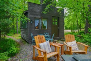 En stuga med två trästolar i Adirondack-stil och ett bord utanför, omgiven av tät grönska.