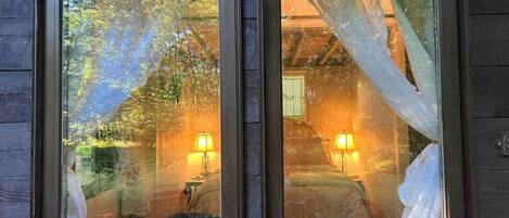 Two windows with curtains, each reflecting a room with a bed and a lamp.