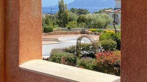 A view from a window with a stone wall and wooden beam, overlooking a landscaped area with a circular water feature, trees, and distant mountains. - Stunning Apartment With Pool in Olbia, Sardinia (Olbia)