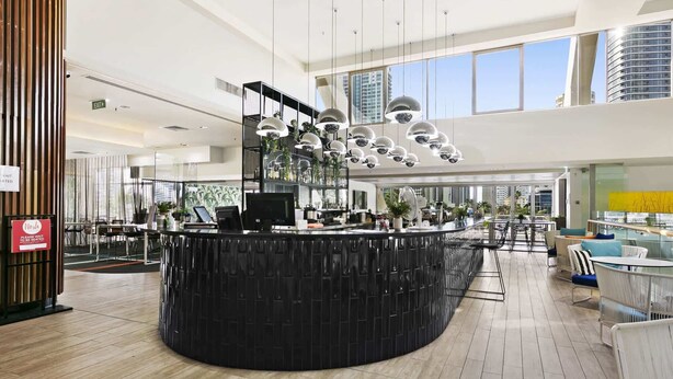 A modern hotel lobby with a central reception desk, multiple seating areas, and large windows offering city views. - Ocean View 2-Bed Studio In the Heart of Surfers (Surfers Paradise)
