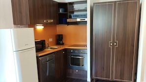 Eine kompakte Küche mit Kühlschrank, Mikrowelle, Kaffeemaschine und Backofen. Ein Geschirrspüler und ein Schrank mit Geschirrgestell sind ebenfalls vorhanden. Der Boden ist mit Fliesen ausgelegt, und die Wände sind weiß gestrichen.