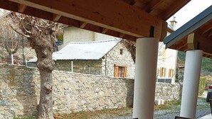A covered outdoor seating area with a round table and chairs, stone walls, and a tree in the background. - 2nd floor, swimming pool, hammam, terrace, parking, tv, 50m², Pra Loup (Jausiers)