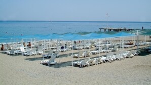 A beach with rows of white sun loungers and umbrellas, a pier extending into the water, and a clear sky. - UK Hotels Kiriş (Kemer)