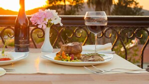 A table with a bottle of wine, a glass of wine, a plate of food, and a vase of flowers on a balcony with a view of trees and a sunset. - UK Hotels Kiriş (Kemer)
