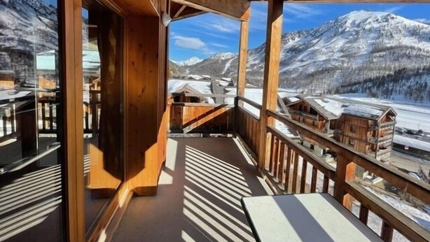 A wooden balcony with a view of snow-covered mountains and a village below. - Apartment with magnificent view in Le Hameau des Airelles (Montgenèvre)