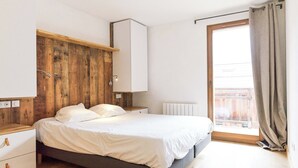A bedroom with a wooden headboard, a white bed, a bedside table with drawers, and a window with curtains. - Apartment with magnificent view in Le Hameau des Airelles (Montgenèvre)