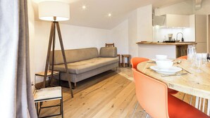 A modern living room with a sofa, a dining table with chairs, a floor lamp, and a kitchen area with a sink and cabinets. - Apartment with magnificent view in Le Hameau des Airelles (Montgenèvre)