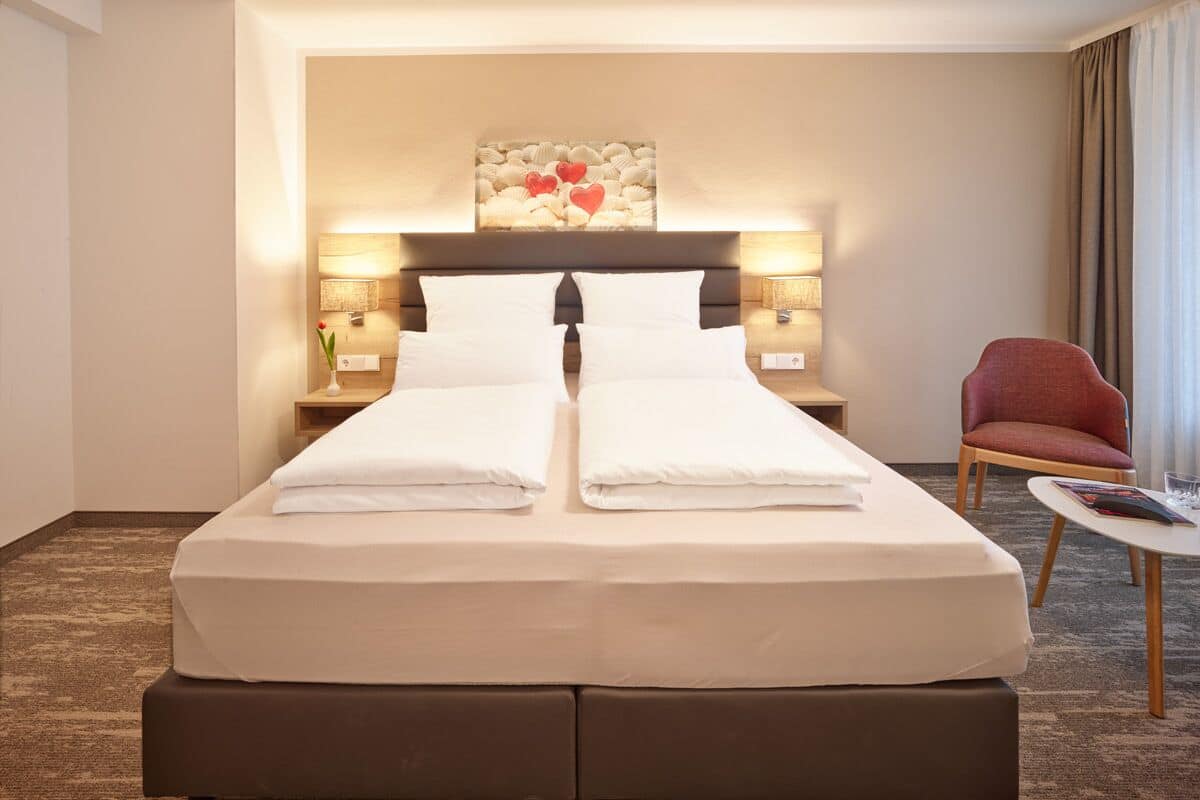 A neatly made double bed with white linens, a headboard with a decorative panel, two bedside lamps, a red chair, and a small table with a magazine.